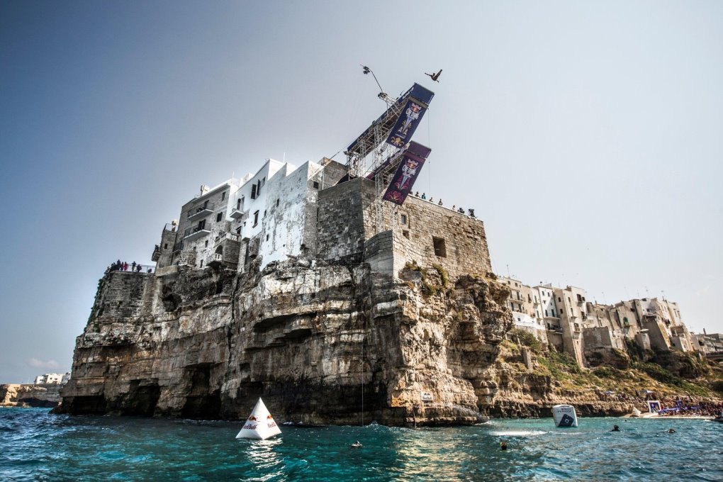 Ein Springer in der Luft von der Plattform des Red Bull Cliff Diving auf den Häusern über der Klippe von Polignano a Mare
