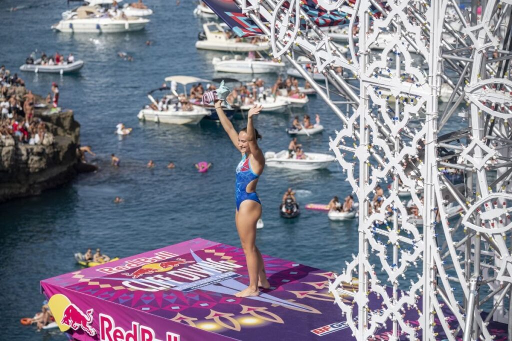 Die Italienerin Elisa Cosetti grüßt das Publikum von der Plattform des Red Bull Cliff Diving in Polignano, unten Boote und Felsen voller Zuschauer