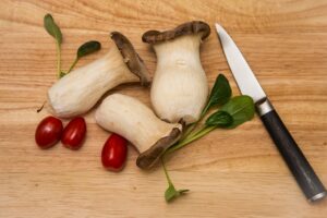 Drei frische Cardoncelli-Pilze auf einem Holzbrett, mit Kirschtomaten und einem Messer