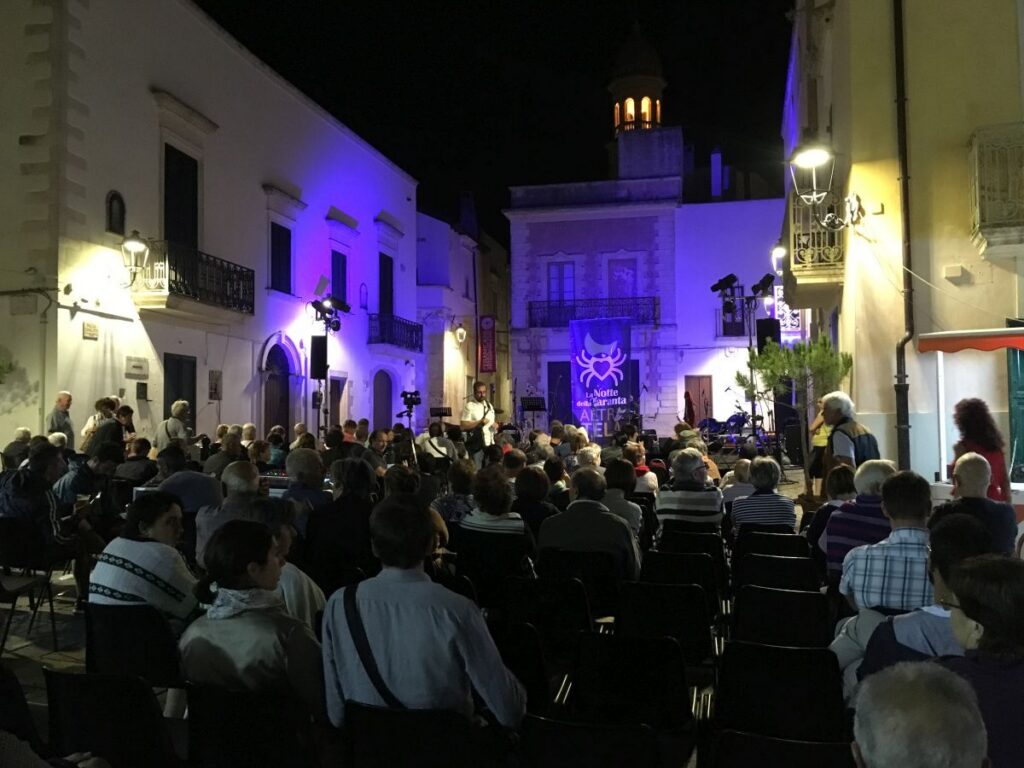 Publikum vor einer Bühne der Notte della Taranta auf einem Dorfplatz im Salento, im Hintergrund das Festivalbanner mit der Spinne