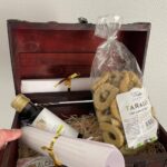 Geöffnete Holztruhe auf Stroh mit einer Tüte Taralli mit Cime di rapa von La Terra di Puglia, einem Fläschchen Olivenöl und einem mit goldenem Band gebundenen Brief