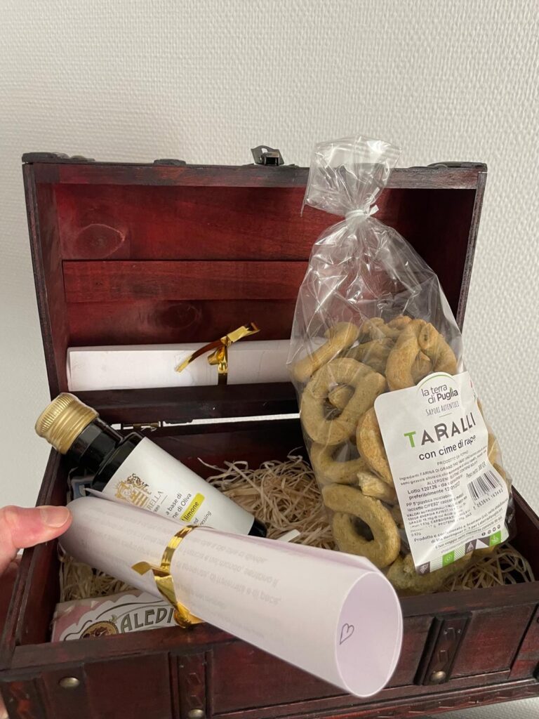 Geöffnete Holztruhe auf Stroh mit einer Tüte Taralli mit Cime di rapa von La Terra di Puglia, einem Fläschchen Olivenöl und einem mit goldenem Band gebundenen Brief