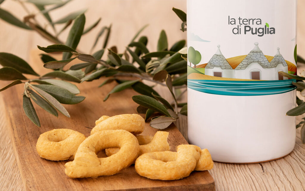 Apulische Taralli auf einem Holzbrett mit Olivenzweigen, daneben ein Ölkrug von La Terra di Puglia